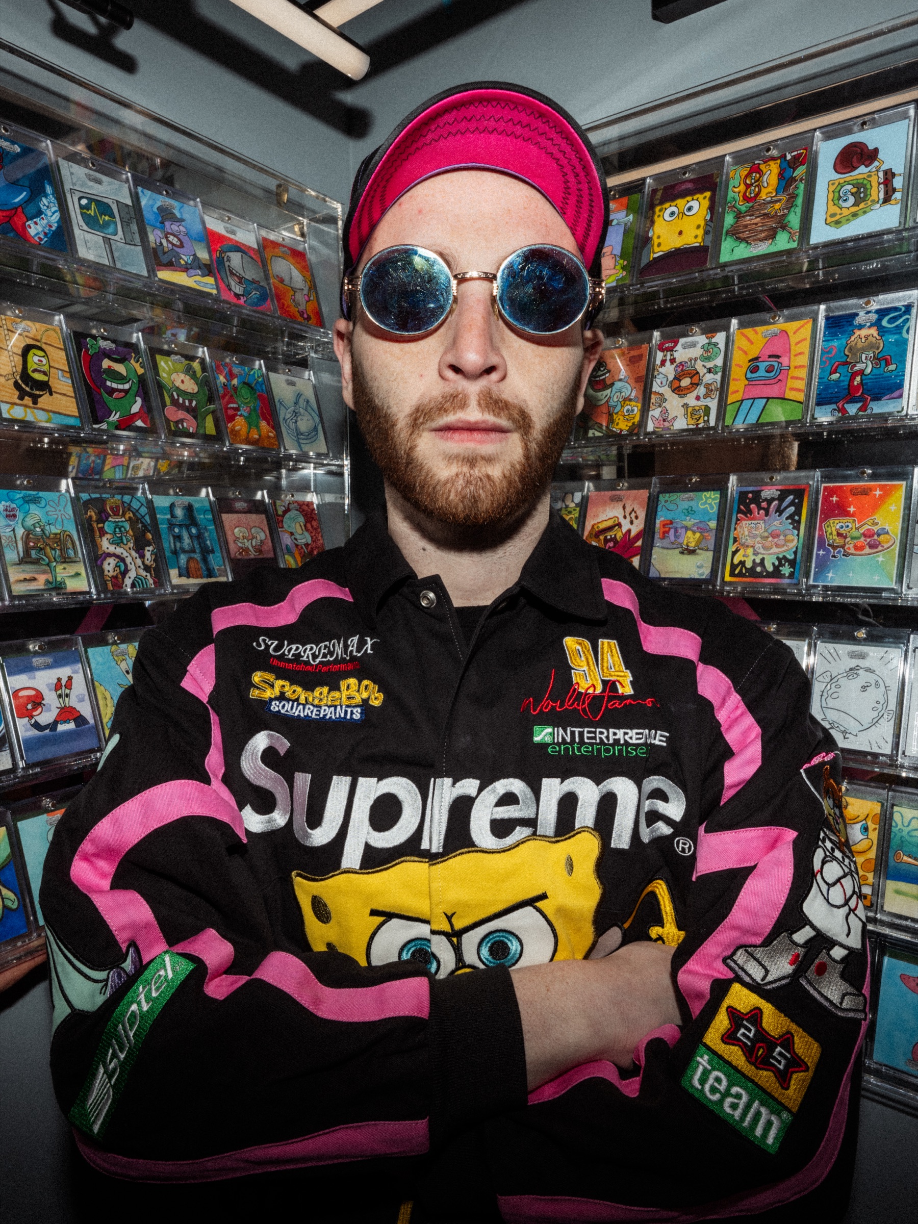 Ari Matlick standing with arms crossed in round sunglasses and a pink and black racing jacket, surrounded by walls of slabbed trading cards.